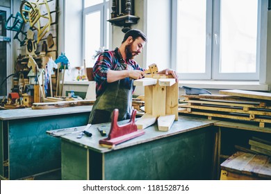 Serious Male Joiner In Apron Using Tools For Making Furniture From Wooden In Workshop, Concentrated Owner Of Manufacturing Business Making Crafts Details By Himself Using Instruments On Working Place