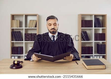 Serious male judge reads legal documents on case sitting at table in his office. Portrait of middle aged man in mantle judge sitting near gavel and court book with folder of documents.