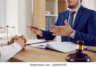 Serious male lawyer explains legal situation and discusses contractual documents with male client. Focus on hands of lawyer sitting at table near judge's gavel and scales of justice. Mid section. - Shutterstock ID 2160995293