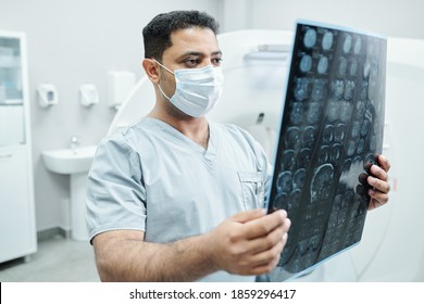 Serious Male Radiologist In Mask And Uniform Looking At X-ray Image Of Patient And Analyzing It Against Environment Of Medical Office