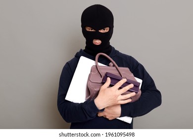 Serious male thief holding bunch of stolen things, laptop, wallet and woman's handbag, robber wearing mask and black turtleneck, burglar isolated over gray background.