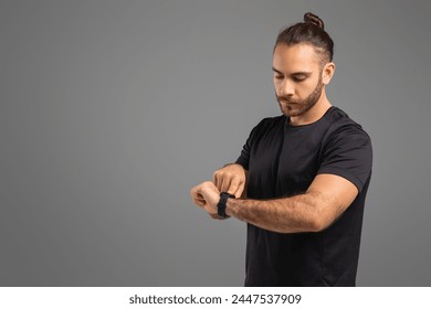 Serious man adjusting a smartwatch on his wrist, wearing a black t-shirt on a grey backdrop, doing workout - Powered by Shutterstock