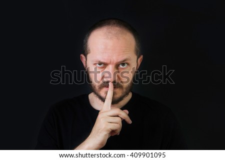     Serious man with beard and mustaches on black background in low key, making silence gesture, pst, shh 