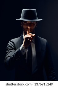 Serious man in black suit and hat keeps finger on lips, making hush gesture and keep conspiracy isolated on dark background. Shh, silence concept                           