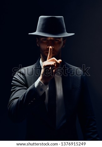 Serious man in black suit and hat keeps finger on lips, making hush gesture and keep conspiracy isolated on dark background. Shh, silence concept                           