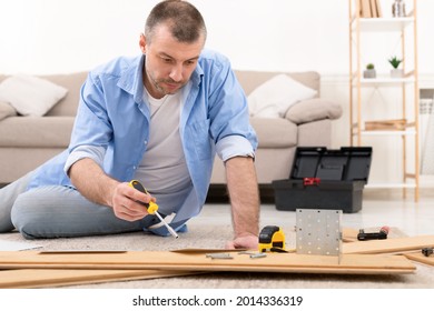 Serious Man Fixing Wooden Cabinet Using Screwdriver And Carpentry Tools Sitting On Floor In Living Room. Mature Male Reading Instruction Furnishing Home, Assembling Furniture With Self-Assembly Kit