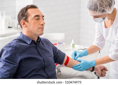 Serious man is having blood test in clinic. Nurse is sticking needle into his vein with concentration