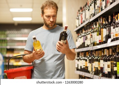 Serious man  holding two bottles of red and white wine and red basket. Client thinking and choosing beverages. Buyer standing in section with alcoholic beverages in supermarket. - Powered by Shutterstock