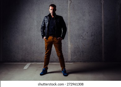 Serious Man In Leather Jacket Looking At Camera, Standing In Empty Garage Against Concrete Wall