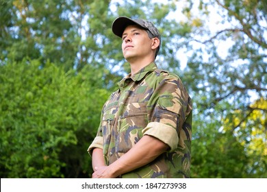 Serious Man In Military Camouflage Uniform Standing In Park, Looking Away. Green Trees Branches In Background. Low Angle Military Man Or Guard Concept