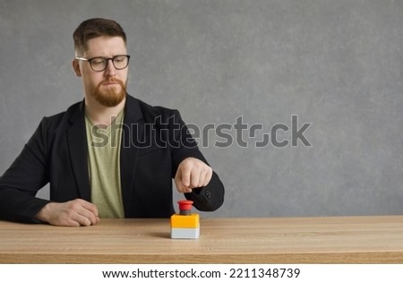 Serious man pushes big red button. Young hipster guy or businessman in glasses hits red alert button on table. Male entrepreneur sitting at desk presses activation button to get access to something