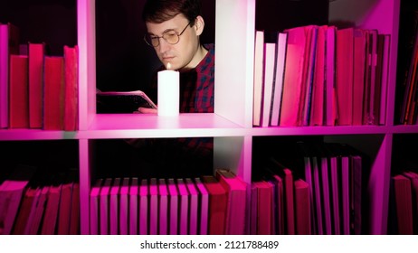 Serious man reading book near candle in bookcase. Male bookworm in eyeglasses reading book near burning candle placed on shelf of wooden bookcase with various literature in pink illumination