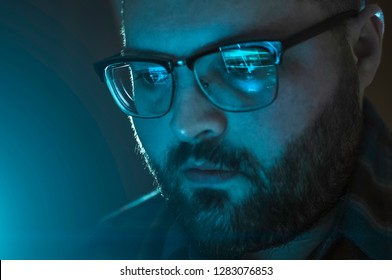 Serious man with reflection of laptop screen with website data in glasses working late, close up. Information analytic or internet marketing or coder developer portrait in blue tones