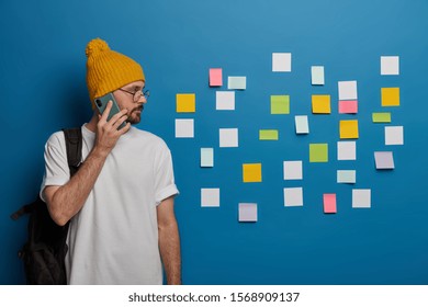 Serious man with satchel on back, has phone talk, concentrated aside on blue wall with many colorful paper notes, discusses and plans work or exam preparation, wears yellow hat and white t shirt