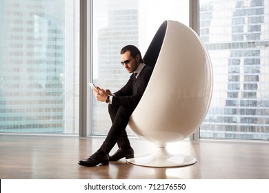 Serious man in sunglasses looking at electronic tablet. Handsome businessman sitting in futuristic egg chair, working on gadget. Shopping online, researching stock market, managing business concept.