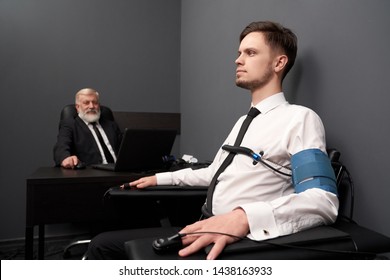 Serious Man Telling Truth During Lie Test On Detector. Young Man In White Shirt Sitting On Chair With Pulse Indicator And Answering Questions. Elder Man Checking Result On Computer At Background.