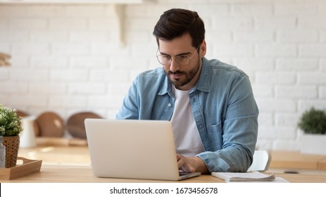 Serious man wearing glasses working on laptop online, sitting at table in kitchen, looking at computer screen, focused male using internet banking service, writing email, searching information