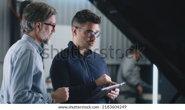 Serious\
manager in a suit and glasses communicating with a man and\
diagnosing a car using a gadget in a car\
service