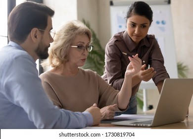 Serious Mature 60 Year Old Businesswoman Using Laptop With Young Indian Female And Businessman Mentor Signing Contract In Office At Meeting. Confident Diverse Woman Managers Talking About New Business