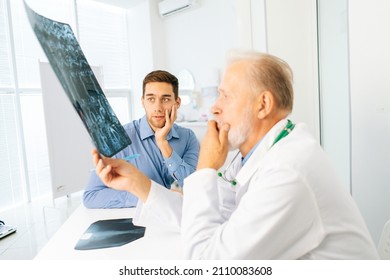 Serious Mature Adult Male Physician Consult Frustrated Young Man Patient Giving Bad News Explaining Results Of MRI Image. Unhappy Scared Young Man Listening To Bad News Sitting In Doctor Office.