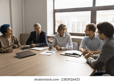 Serious mature business leader woman talking to diverse colleagues at office table, explaining tasks to employees. Multiethnic team meeting for conversation, collaboration, teamwork - Powered by Shutterstock