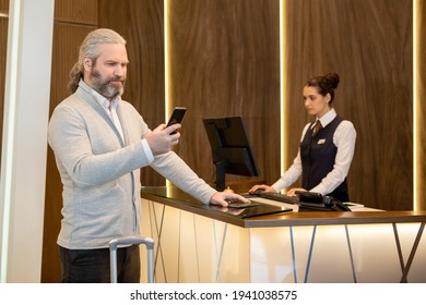 Serious Mature Businessman In Smart Casualwear Looking At Smartphone Screen While Standing By Reception Counter In The Hall Of Hotel