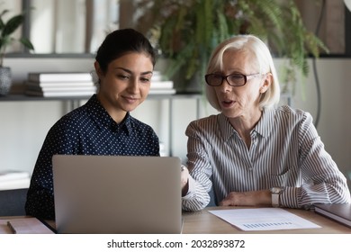 Serious Mature Businesswoman Mentor Coach In Glasses Teaching Indian Woman Intern Student, Using Laptop, Pointing At Screen, Two Diverse Colleagues Working On Online Project Together, Discussing