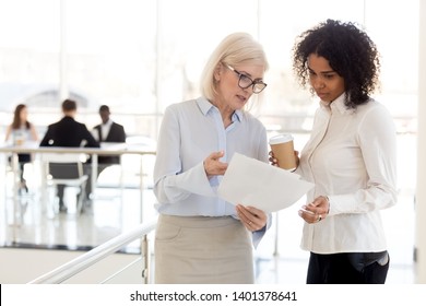 Serious mature businesswoman show document to African American female colleague during break, middle-aged boss explaining task, point at mistake to subordinate, diverse employees discuss paperwork