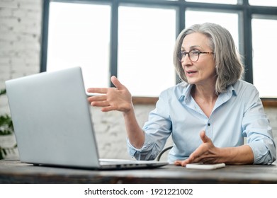 Serious Mature Businesswoman Sitting At The Desk, Looking At Webcam And Talking With Client Online, Elder Female Office Worker Complaining Gesturing Or Arguing On A Virtual Meeting Via Laptop