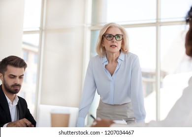 Serious mature businesswoman stand talk with employees at office meeting, middle aged female boss discuss project or strategy with workers at office briefing, confident woman negotiate with colleagues