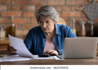 Serious Mature Caucasian 60s Woman Sit At Table Calculate Household Finances Paying Bills On Laptop Online. Focused Senior Grey-haired Grandmother Manage Family Home Budget, Read Paper Bank Notice