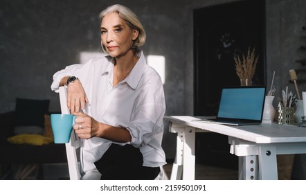 Serious Mature Caucasian Female Entrepreneur With Cup Of Tea Sitting On Chair And Looking At Camera. Small Business And Entrepreneurship. Modern Successful Woman At Dark Art Studio With Sunny Lighting