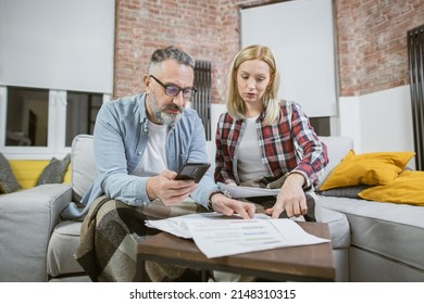 Serious Mature Couple With Modern Smartphone And Paper Calculating Domestic Finances While Sitting Together On Couch. Concept Of People And Home Budgeting.