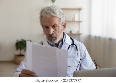 Serious Mature Doctor Reading Medical Document, Working In Office, Focused Senior Therapist Physician Gp Wearing White Uniform Sitting At Desk In Hospital, Analyzing Patient Diagnosis Or Report