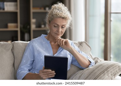 Serious mature female using digital tablet, sit on sofa with device, read on-line news, learning new application looks concerned, due to lack of understanding. Older gen and modern wireless tech usage - Powered by Shutterstock