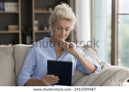 Serious mature female using digital tablet, sit on sofa with device, read on-line news, learning new application looks concerned, due to lack of understanding. Older gen and modern wireless tech usage