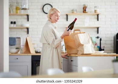 Serious Mature Lady With Short Blond Hair Standing At Kitchen Counter And Reading Label Of Wine Bottle While Unpacking Paper Bags In Kitchen