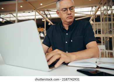 Serious Mature Male Executive Wearing Eyeglasses Looking At Diary While Doing Research Over Laptop On Desk In Office