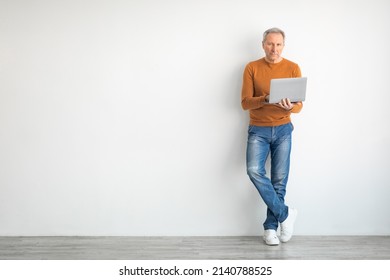 Serious Mature Man Holding Using Laptop Standing Leaning On White Wall, Posing Looking At Camera. Adult Male Typing On Keyboard Isolated On Studio Background, Full Body Length, Free Copy Space