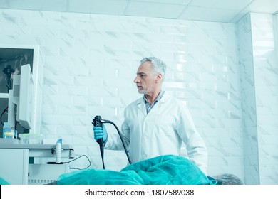 Serious mature medical worker holding an endoscope and looking at the screen while standing next to his patient
