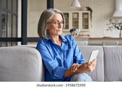 Serious Mature Middle Age Senior Woman At Home On Couch Holding Mobile Cellphone, Reading News Or Watching Online Learning Class Having Video Call Using Mobile Application.