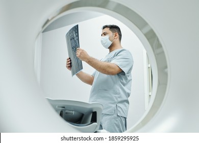 Serious Mature Mixed-race Radiologist In Mask And Uniform Looking At X-ray Image Of Patient Head In Medical Office Environment In Clinics