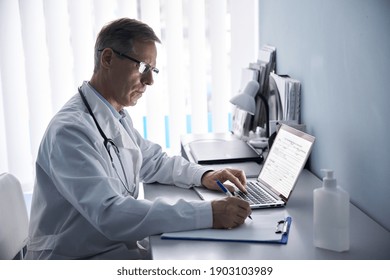 Serious mature old doctor physician making medical notes in notebook using laptop in hospital office. Senior middle aged male practitioner working on computer and writing sitting at desk.