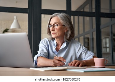 Serious mature older adult woman watching training webinar on laptop working from home or in office. 60s middle aged businesswoman taking notes while using computer technology sitting at table. - Shutterstock ID 1827080276
