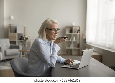 Serious mature older business lady recording voice message on mobile phone at work table with laptop, speaking at cellphone, giving audio command for search on Internet - Powered by Shutterstock