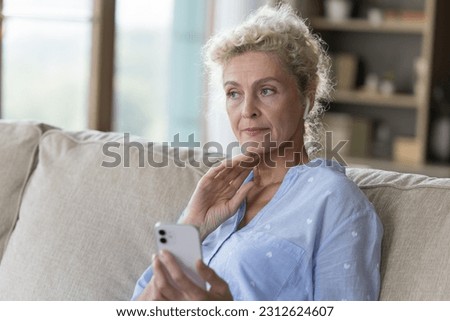 Serious mature senior woman lady holding mobile phone, sitting on home couch, looking away, touching chin, thinking, using online app for communication, Internet connection, feeling concerned