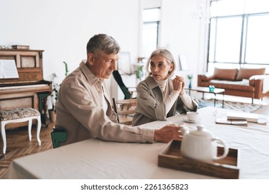 Serious mature woman in casual clothes looking at husband having conversation during coffee break near table in morning at home - Powered by Shutterstock