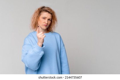 Serious middle age woman with curly blonde hair shaking her finger, scolding or telling off someone, standing in trendy blue sweatshirt over light grey background