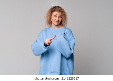 Serious middle age woman with curly blonde hair shakes her finger threateningly, scolding or telling off someone, standing in trendy blue sweatshirt over light grey background