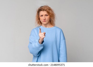 Serious middle age woman with curly blonde hair is warning, shakes her finger threateningly, standing in trendy blue sweatshirt over light grey background